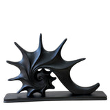 A black abstract sculpture shaped like a stylized spiral shell or nautilus, mounted on a rectangular black base against a plain white background.