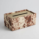 A rectangular tissue box cover with a brushed gold metal slot on top and a brown and beige splattered patterned Lather