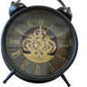A black twin-bell alarm clock with a transparent glass face showing an ornate gold inner gear mechanism and gold Roman numerals.