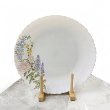 A white plate with a scalloped rim, decorated with a floral pattern of pink, purple, and yellow flowers with green leaves on the left side, displayed on a small wooden stand.