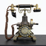 An antique-style rotary telephone with a dark gray and brass/gold metal body, featuring a coiled brown cord, a black handle, and a circular dial face with decorative brass accents, displayed on a dark tabletop.