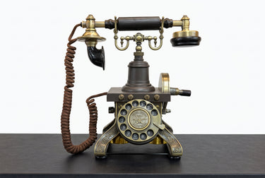 An antique-style rotary telephone with a dark gray and brass/gold metal body, featuring a coiled brown cord, a black handle, and a circular dial face with decorative brass accents, displayed on a dark tabletop.